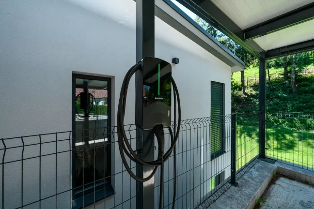 Elektroauto-Ladestation am Pfosten des anthrazitfarbenen Carports an verputzter Fassade.