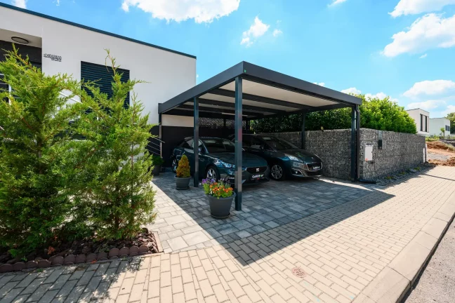 Zwei Fahrzeuge unter anthrazitfarbenem Stahl-Flachdach-Carport am Wohnhaus.