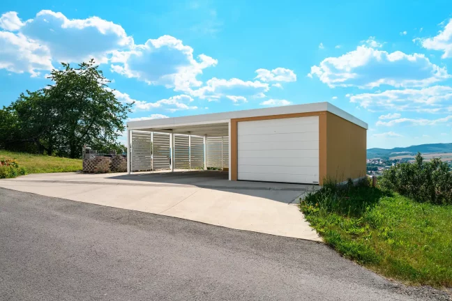 Garage mit Ziegelputz, Carport und weißem Sektionaltor an einem Hang.