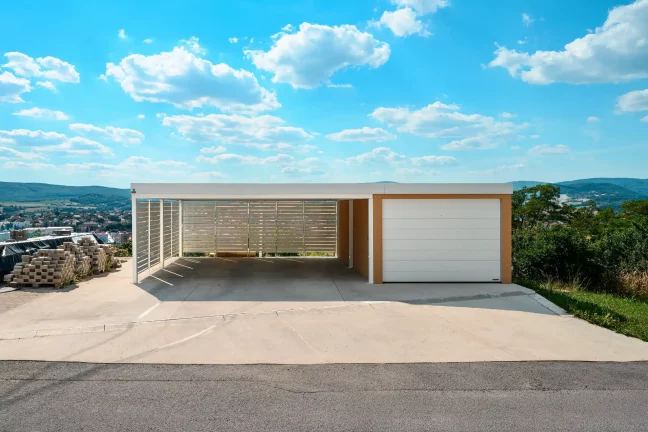 Offener Carport mit Metallverkleidung und Garage mit Ziegelputz auf erhöhtem Grundstück.