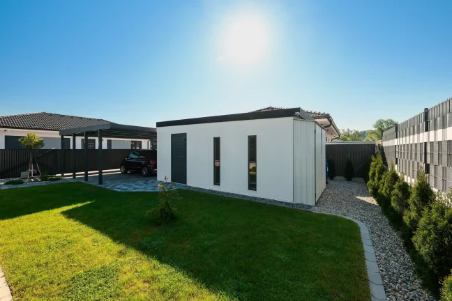 Weiße Garage mit anthrazitfarbenem Pultdach und gepflegtem grünen Garten am Zaun