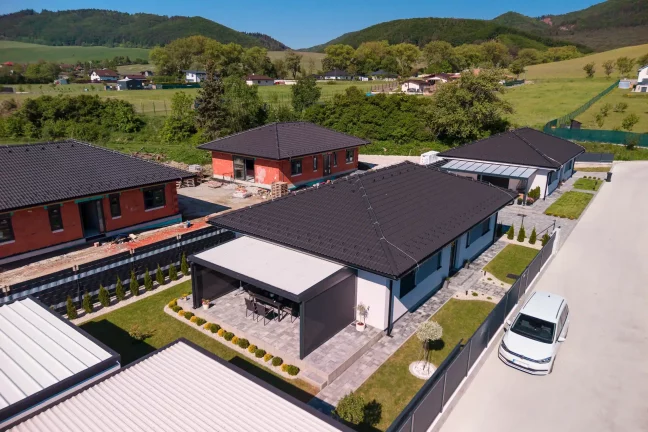 Luftaufnahme einer Pergola mit Flachdach neben einem weißen Einfamilienhaus, grüne Umgebung mit Bergen