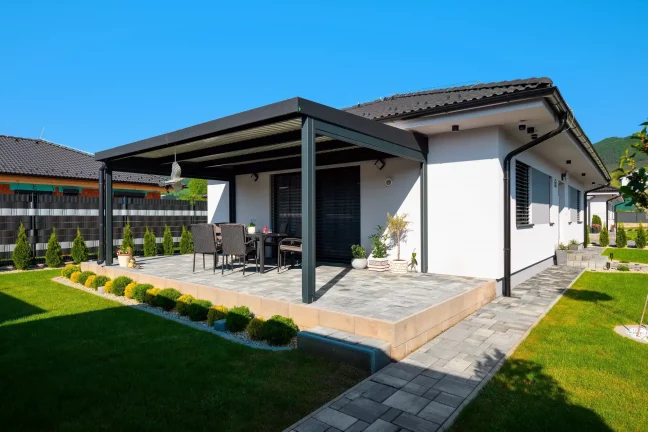 Anthrazitfarbene Pergola neben einem weißen Haus mit gepflasterter Terrasse und Gartenmöbeln, blauer Himmel