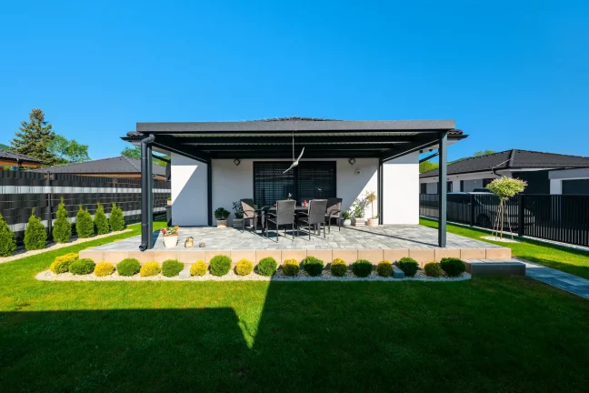 Anthrazitfarbene Pergola mit Pultdach, Gartenmöbel und Ziersträucher rundherum