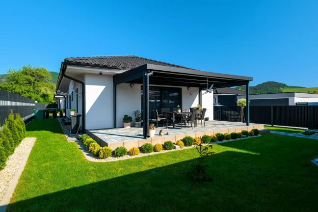 Pergola mit anthrazitfarbener Konstruktion neben einem weißen Haus, grüner Rasen
