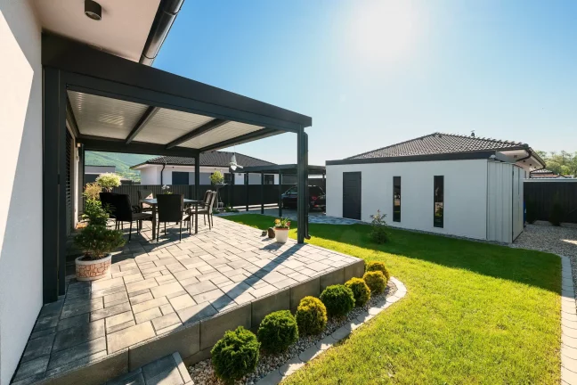 Seitenansicht einer anthrazitfarbenen Pergola mit Terrassenmöbeln und gepflegtem Garten am Haus