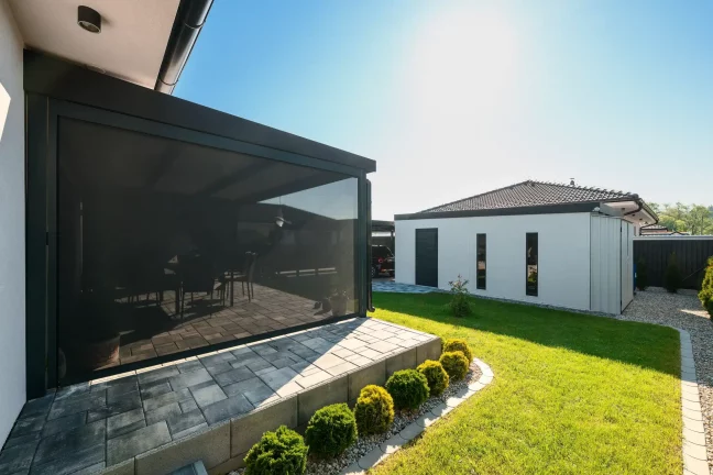 Anthrazitfarbene Pergola mit heruntergelassenem Rollo neben einem weißen Haus, gepflasterte Terrasse und grüner Rasen