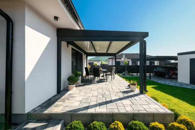 Anthrazitfarbene Pergola mit Lamellendach neben einem weißen Haus, gepflasterte Terrasse mit Gartenmöbeln