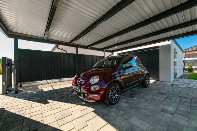 Carport mit flachem Pultdach und anthrazitfarbenen Stützen neben einem weißen Haus, grüner Rasen