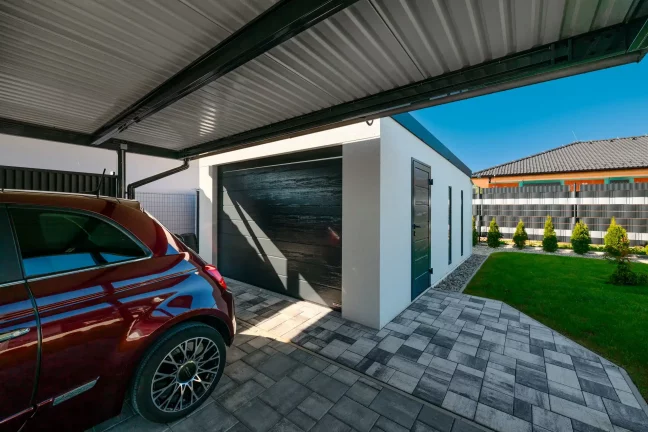Blick aus dem Inneren des Carports auf ein rotes Auto, anthrazitfarbenes Pultdach und weiße Garage im Hintergrund