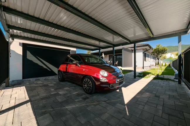 Rotes Auto unter einem Carport mit Pultdach, Seitenansicht, gepflasterte Fläche und grüner Garten