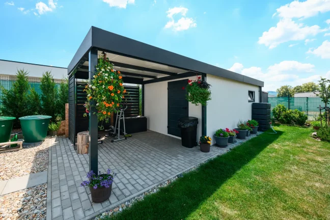 Blumengeschmückter Garten mit weißem Gartenhaus und anthrazitfarbenem Carport.
