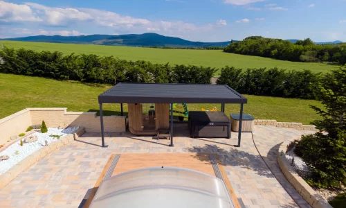 Anthrazitfarbene Pergola mit Gartenmöbeln neben einem Pool mit transparenter Abdeckung, grüne Hügel im Hintergrund