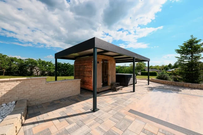 Freistehende anthrazitfarbene Pergola mit Flachdach auf einer gepflasterten Terrasse, grüner Garten im Hintergrund