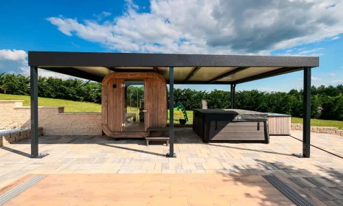 Anthrazitfarbene Pergola mit Flachdach neben einer Fasssauna auf einer gepflasterten Terrasse, blauer Himmel