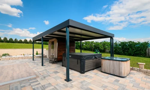 Anthrazitfarbene Pergola mit Whirlpool darunter auf einer gepflasterten Terrasse, grüner Garten und blauer Himmel