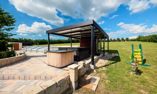 Anthrazitfarbene Pergola mit Whirlpool und Kinderspielplatz im Hintergrund, gepflasterte Terrasse am Garten