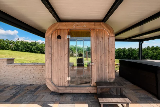 Blick aus dem Inneren einer anthrazitfarbenen Pergola auf eine Fasssauna mit Holztür und Whirlpool