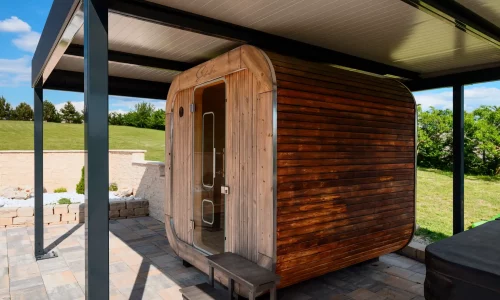 Fasssauna aus dunklem Holz unter einer anthrazitfarbenen Pergola, gepflasterte Fläche und grüner Rasen