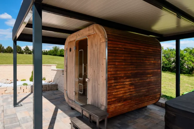Fasssauna aus dunklem Holz unter einer anthrazitfarbenen Pergola, gepflasterte Fläche und grüner Rasen