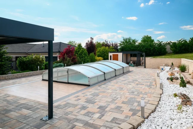 Anthrazitfarbene Überdachung am Pool mit transparenter Abdeckung, gepflasterte Fläche und Zierpflanzen