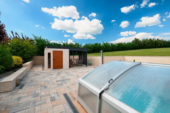 Blick auf einen Pool mit transparenter Abdeckung und ein Gartenhaus mit braunen Türen, gepflasterte Terrasse