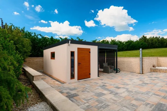 Gartenhaus mit braunen Türen auf einer gepflasterten Terrasse, blauer Himmel