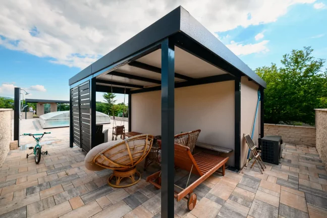 Anthrazitfarbene Pergola neben einem Gartenhaus mit Türen, Fahrrad und Gartenmöbel darunter