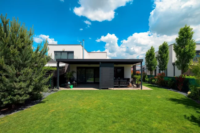 Anthrazitfarbene Pergola mit Flachdach neben einem weißen Haus, weitläufiger grüner Rasen und Bäume