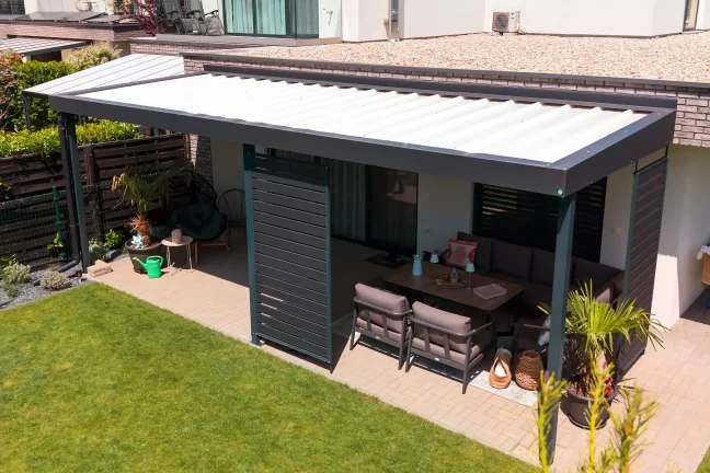 Pergola mit Gartenmöbeln an einer dunklen Fassade, gepflasterte Terrasse