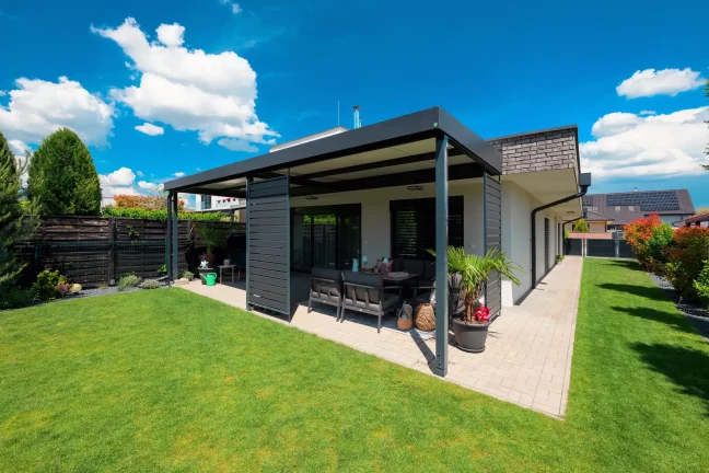 Anthrazitfarbene Pergola mit Gartenmöbeln, Seitenansicht auf den grünen Garten