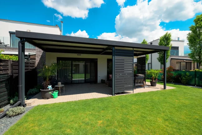 Freistehende anthrazitfarbene Pergola mit Flachdach im grünen Garten, blauer Himmel mit Wolken