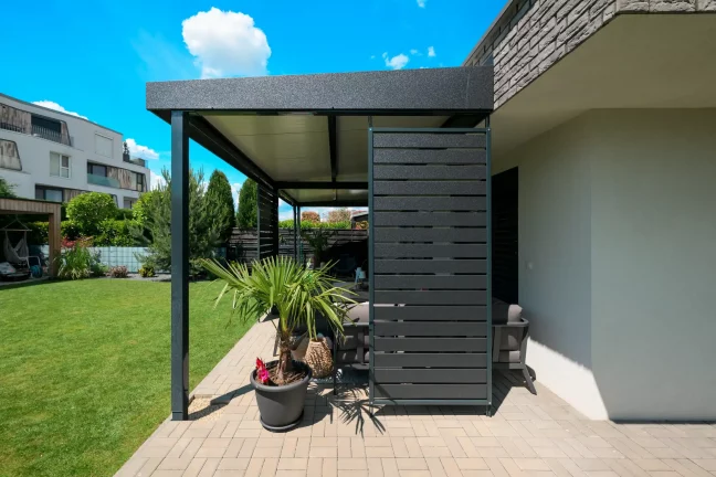 Anthrazitfarbene Pergola mit Seitenverkleidung neben einem Backsteinhaus, dekorative Pflanzen auf der Terrasse