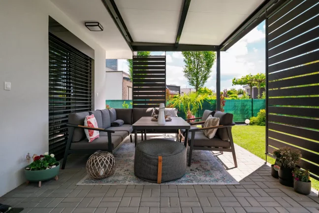 Blick aus dem Inneren einer anthrazitfarbenen Pergola auf Gartenmöbel und Seitenelemente